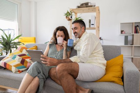 Téléchargez les photos : Couple marié adulte regardant leur tablette tout en buvant avec leur tasse assis sur leur canapé dans le salon partageant le temps de la famille - en image libre de droit