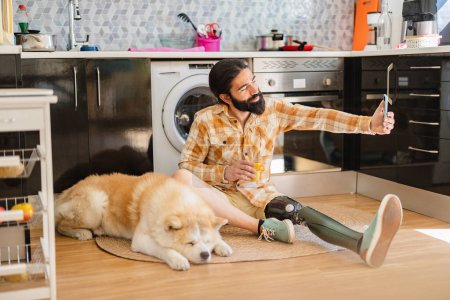 Téléchargez les photos : Homme avec la jambe prothétique assis sur le sol de la cuisine avec le mobile et son chien couché - en image libre de droit