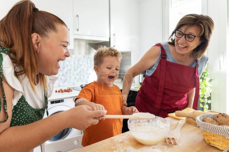Téléchargez les photos : Plan moyen de grand-mère, fille et petit-fils passer un bon moment dans la cuisine tout en riant et en mélangeant les ingrédients - en image libre de droit