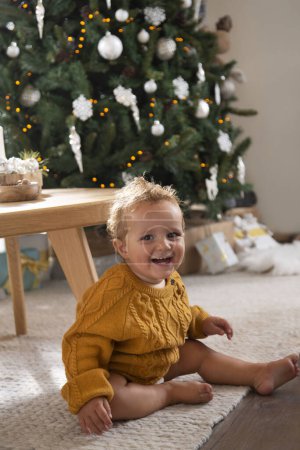 Foto de Niño sentado en el suelo después de arrastrarse por su sala de estar decorada con decoraciones de Navidad y riendo en voz alta frente al árbol de Navidad - Imagen libre de derechos