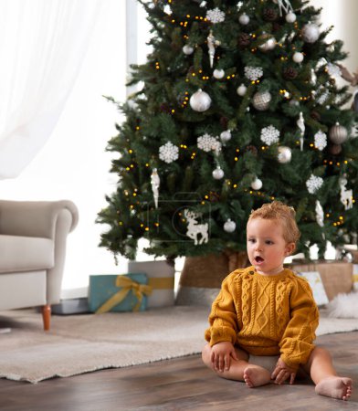 Téléchargez les photos : Bébé garçon assis sur le sol en face de l'arbre de Noël dans le salon de sa maison décorée de lumières et de guirlandes. Il a une expression de surprise avec la bouche ouverte attendant ses cadeaux de Noël - en image libre de droit