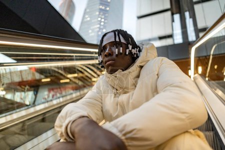Téléchargez les photos : Sérieux jeune homme noir branché assis sur l'escalator dans un centre commercial de la ville - en image libre de droit