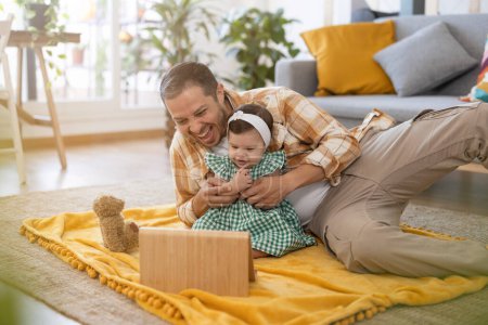 Foto de Papá latino en el suelo de su salón jugando con su bebé mientras ven los dibujos en su tableta - Imagen libre de derechos
