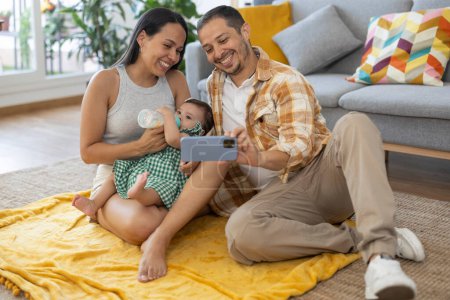 Foto de Latina esposa dando a su bebé un biberón mientras toma una selfie con su marido felizmente en su sala de estar - Imagen libre de derechos