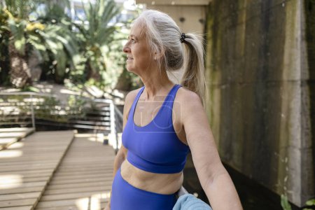Téléchargez les photos : Sportive aux cheveux gris femme âgée se reposant dans le parc après l'entraînement, porte des vêtements de sport bleu serré - en image libre de droit