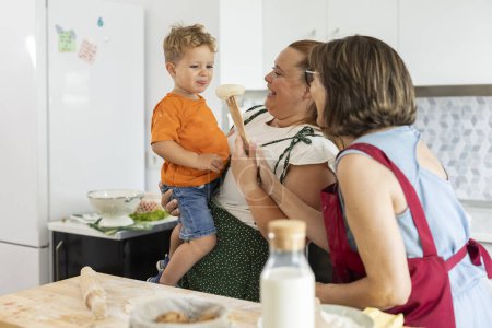Téléchargez les photos : Mère et grand-mère avec leurs tabliers découvrant des saveurs avec leur fils et petit-fils dans la cuisine - en image libre de droit
