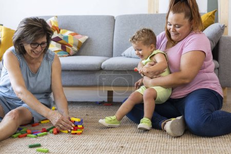 Foto de Familia juega en el suelo de su casa mientras la madre sostiene a su hijo para evitar derribar los bloques de juego - Imagen libre de derechos