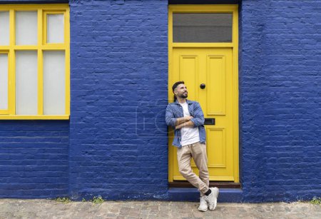 Foto de Joven latino espera con los brazos cruzados su cita apoyada en una pared azul con una puerta amarilla típica de Londres. - Imagen libre de derechos