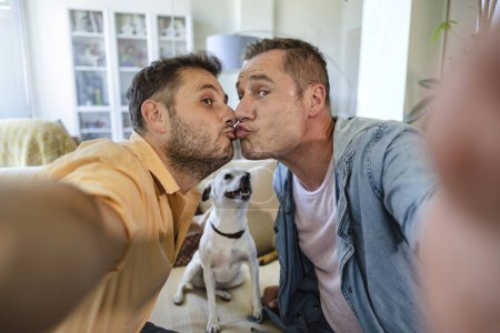 Téléchargez les photos : Couple gay prenant un selfie pour la Saint-Valentin tout en embrassant. Leur chien les regarde. - en image libre de droit