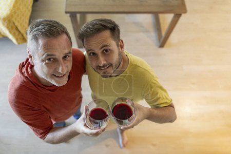 Foto de Fotografía aérea de pareja gay adulta brindando con su copa de vino mientras mira a la cámara - Imagen libre de derechos