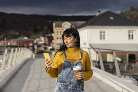 Foto de Mujer adulta joven mirando su móvil mientras camina y lleva café en la mano - Imagen libre de derechos