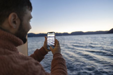Photo for Young adult tourist man taking a photo at dusk with his smartphone on a boat on the sea - Royalty Free Image