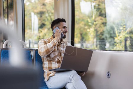 Foto de Sonriente hombre de negocios trabajando en su computadora en el tren mientras habla por teléfono - Imagen libre de derechos