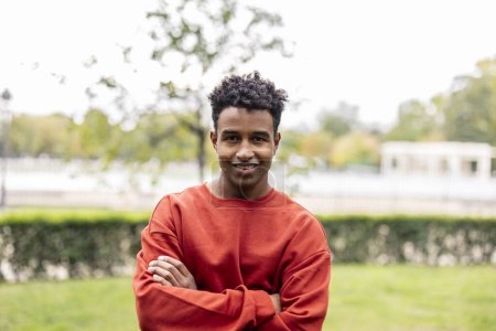 Téléchargez les photos : Portrait de jeune homme afro-américain adulte avec les bras croisés et un pull rouge regardant la caméra tout en souriant - en image libre de droit