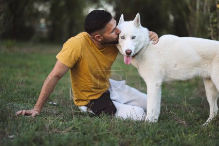 Foto de Atractivo hombre sentado con su perro en la hierba mientras lo besa - Imagen libre de derechos
