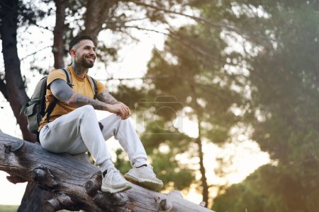 Photo for Smiling tattooed model man resting sitting on tree trunk in forest at sunset with back light - Royalty Free Image