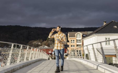 Téléchargez les photos : Homme hispanique marchant vers la caméra boire du café et écouter de la musique avec son casque avec la ville en arrière-plan - en image libre de droit