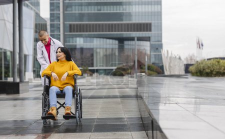 Téléchargez les photos : Une jeune beauté en fauteuil roulant est prise par une infirmière pour une promenade dans la rue près de l'hôpital - en image libre de droit