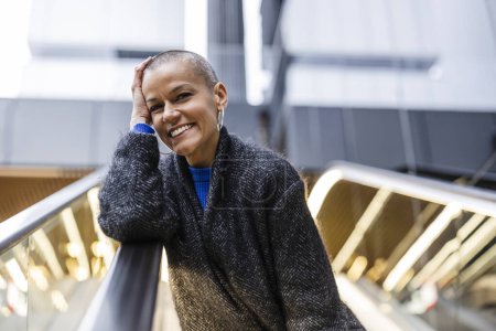 Photo for Bald shaved woman posing very smiling in a shopping center going up escalators and surrounded by lights, she is happy about her therapy in recovering from her breast cancer - Royalty Free Image