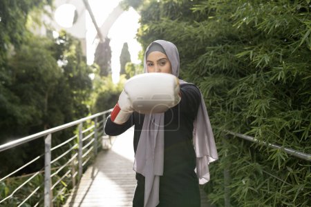 Téléchargez les photos : Jeune femme musulmane voilée jetant un coup de poing à la caméra avec des gants de boxe - en image libre de droit