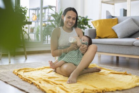 Foto de Madre latina mirando a la cámara sonriendo dando biberón a su bebé sentado en la alfombra - Imagen libre de derechos