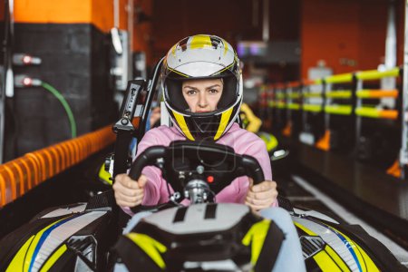 Téléchargez les photos : Femme pilote regardant avec défiance la caméra avec son casque de course tenant le volant de la voiture de karting à la ligne de départ sur le circuit - en image libre de droit