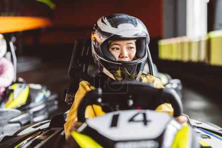 Téléchargez les photos : Jeune asiatique femme kart racer avec casque tenant le volant dans sa voiture de course sur le circuit récréatif - en image libre de droit
