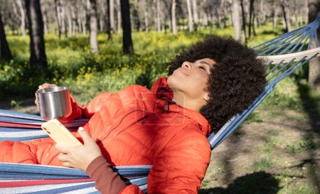 Foto de Hermosa mujer afroamericana acostada descansando en una hamaca en la naturaleza ajardinada con el teléfono móvil en la mano - concepto de desconexión digital - Imagen libre de derechos