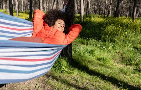 Téléchargez les photos : Jeune femme afro-américaine en hamac se reposant heureux et souriant avec les yeux fermés profitant d'un beau moment de la vie sauvage au coucher du soleil - en image libre de droit