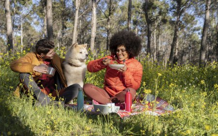 Téléchargez les photos : Couple aimant multiethnique déjeuner en pleine nature avec leur chien - rendez-vous dans la forêt - en image libre de droit