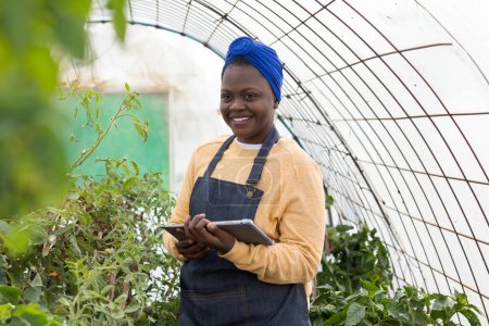 Cheerful young farmer uses a tablet to manage greenhouse operations, amidst thriving plants.