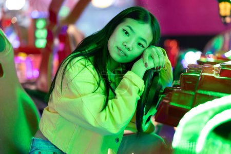 Foto de Retrato de joven mujer étnica asiática apoyada en el volante de una máquina de juego arcade virtual con luces de neón mirando a la cámara - Imagen libre de derechos