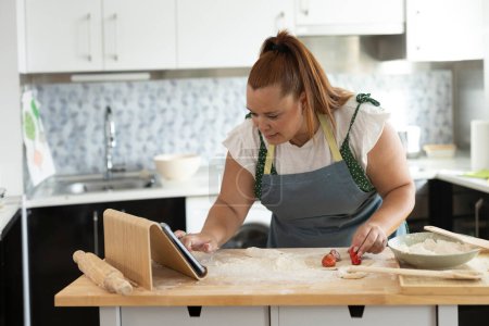 Téléchargez les photos : Curvy femme toucher l'écran de sa tablette numérique tout en cuisinant pour apprendre de la recette en ligne tutoriel - en image libre de droit