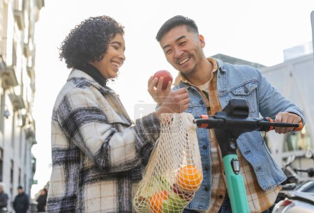 Téléchargez les photos : Couple multiracial achète des aliments sains ensemble dans des sacs de filet respectueux de l'environnement sur scooter électrique - en image libre de droit