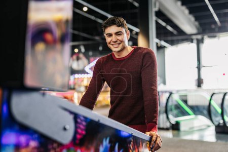 Retrato de un joven guapo sonriente apoyado en una máquina de pinball en una sala de juegos arcade