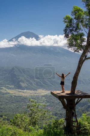 Foto de Vista trasera turista femenina disfrutando de la naturaleza mirando las montañas y el volcán Agung mientras está de pie en el lugar de la foto en el árbol. Bali, Indonesia. Concepto de viaje - Imagen libre de derechos