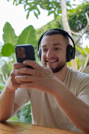 Foto de Joven hombre freelancer creativo feliz en ropa casual trabajando en el espacio de coworking con auriculares en el ordenador portátil. Trabajo remoto en la isla tropical de Bali, reubicación - Imagen libre de derechos
