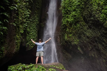 Photo for Back view of a man standing by waterfalls with arms outstretched. Man at waterfall raising his hands in feeling closer to nature. Man at the waterfall. Travel to Bali - Royalty Free Image