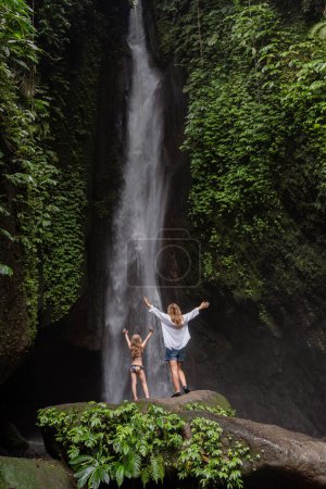 Photo for Family at a tropical waterfall. A woman and child at a beautiful waterfall. Vacation on a tropical island. Traveling couple at the waterfall. Family on vacation in Bali - Royalty Free Image