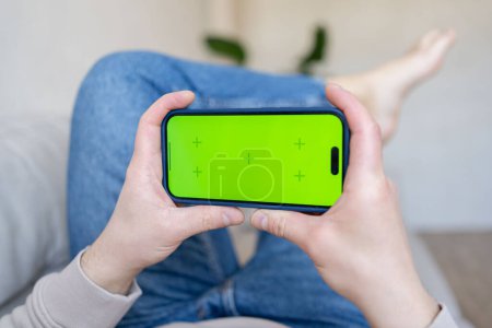 Photo for Man at home lying on a couch using smartphone with green mock-up screen - Royalty Free Image