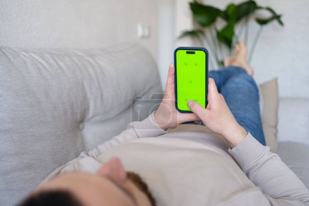 Photo for Man at home lying on a couch using smartphone with green mock-up screen - Royalty Free Image