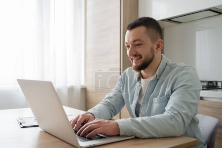 Foto de Retrato de un joven sonriente usando un portátil sentado en el escritorio, escribiendo en un cuaderno. Chico alegre navegar por Internet, viendo webinar estudiar en línea, mirando la pantalla de la PC en casa. - Imagen libre de derechos