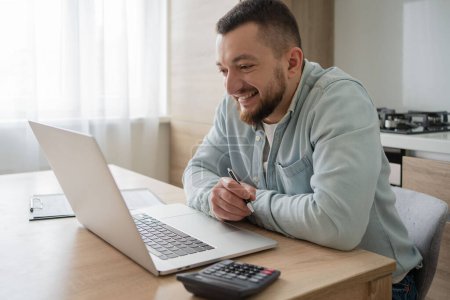 Téléchargez les photos : Portrait d'un jeune homme souriant utilisant un ordinateur portable assis au bureau, écrivant dans un carnet. Gai gars naviguant sur Internet, regarder webinaire étudiant en ligne, regardant l'écran PC à la maison. - en image libre de droit