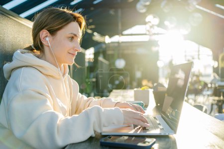 Photo for Young beautiful happy woman in headphones is listening music on laptop in cafe or co working center. Female freelancer wearing headphones working on the notebook - Royalty Free Image