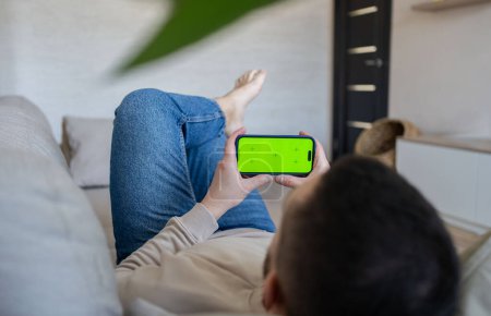 Photo for Man at home resting on a couch using smartphone with green mock-up screen. Guy using mobile phone, internet social networks browsing - Royalty Free Image