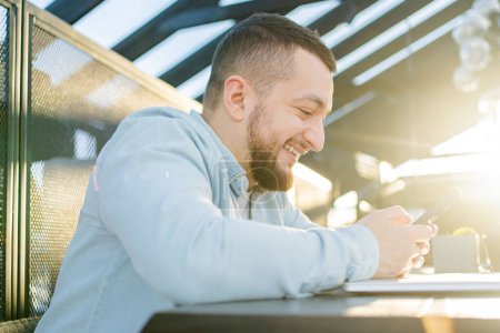 Photo for Cheerful freelancer with beard browsing smartphone and smiling while sitting at table with laptop and notebook in cafe - Royalty Free Image
