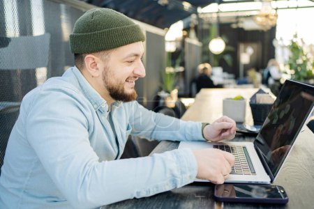 Téléchargez les photos : Jeune homme beau assis dans un café, tapant et utilisant un ordinateur portable. Beau hipster utilisant l'ordinateur au café - en image libre de droit