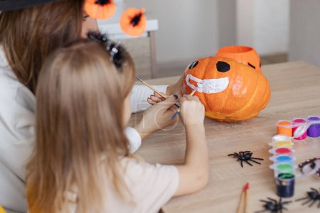 Téléchargez les photos : Petite fille faisant décoration Halloween avec parent à la maison, dessin visage effrayant sur la citrouille et profiter de la préparation des vacances - en image libre de droit