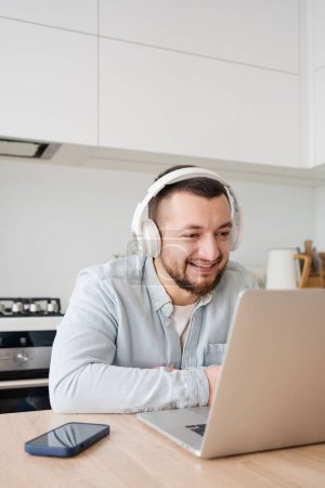 Téléchargez les photos : Jeune homme avec ordinateur portable et écouteurs à l'intérieur à la maison, étudier, travail indépendant - en image libre de droit