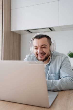 Téléchargez les photos : Jeune homme avec ordinateur portable et écouteurs à l'intérieur à la maison, étudier, travail indépendant - en image libre de droit